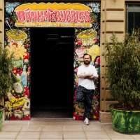 A man stands in front of the entrance to 'Banh Mi & Bubbles', surrounded by colorful graffiti and plants., © Banh Mi & Bubbles Restaurant / Bar A man stands in front of the entrance to 'Banh Mi & Bubbles', surrounded by colorful graffiti and plants., © Banh Mi & Bubbles Restaurant / Bar