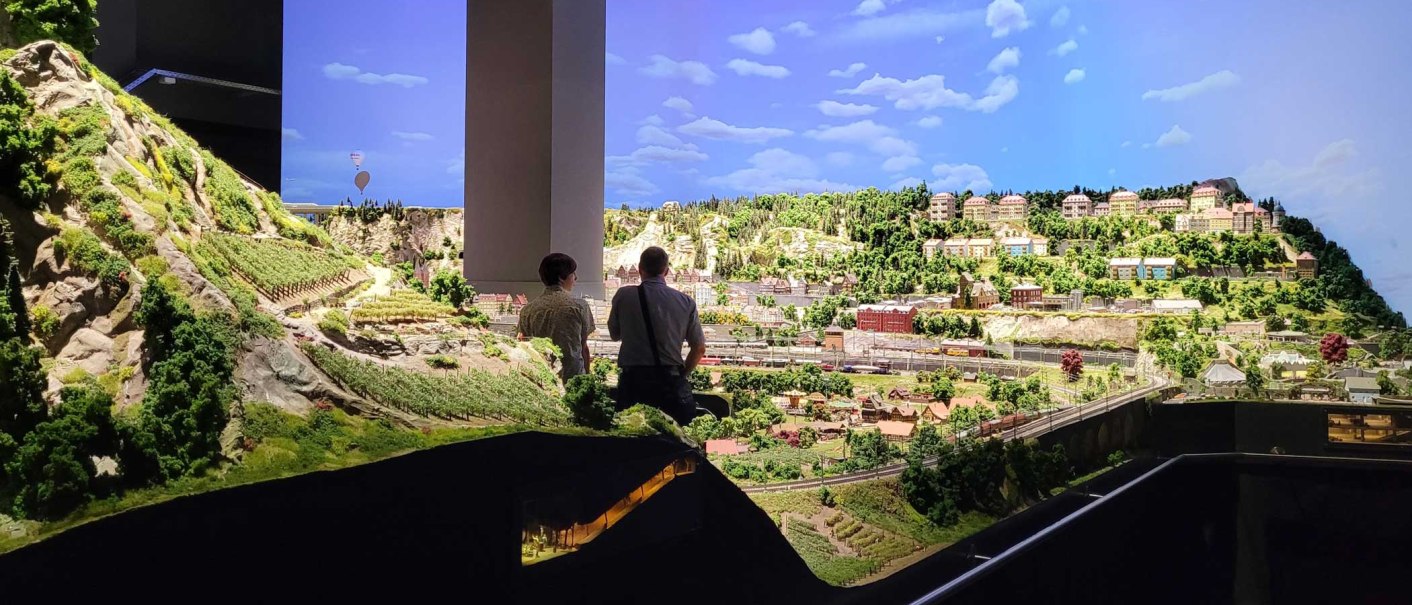 Model railroad layout with detailed landscape and buildings, viewed by two people. The sky is blue with clouds., © Märklineum Model railroad layout with detailed landscape and buildings, viewed by two people. The sky is blue with clouds., © Märklineum