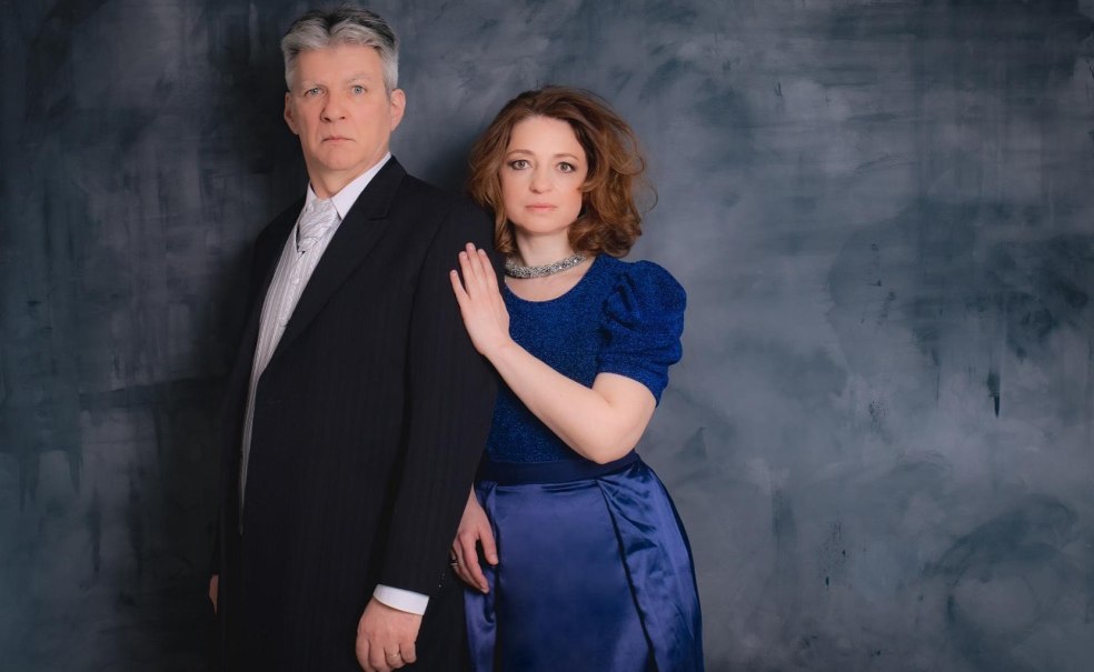 Ein Mann im Anzug und eine Frau in einem blauen Kleid stehen vor einem grauen Hintergrund. Die Frau legt ihre Hand auf die Schulter des Mannes., © Diana Hajdu, Peter Cser