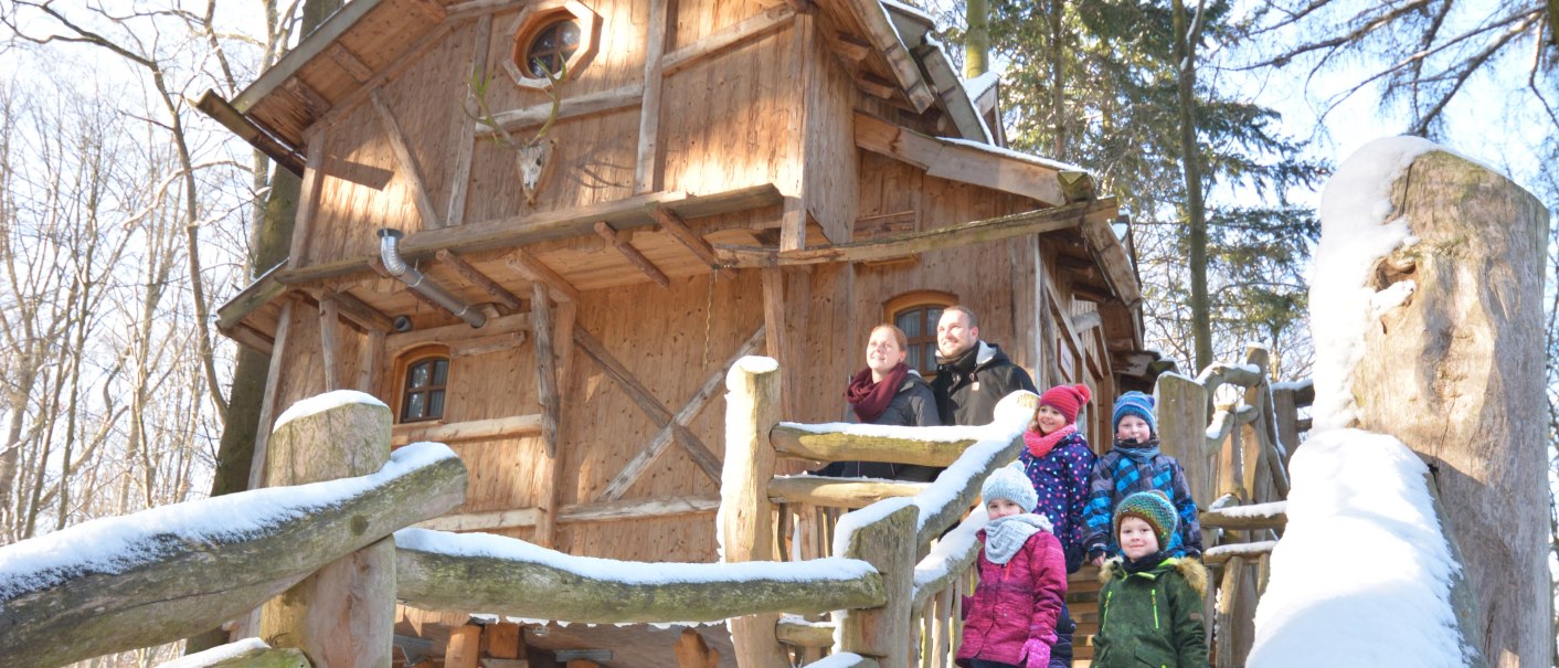 A family is standing on the steps of a snow-covered treehouse in the forest. The wooden house has a rustic look and is surrounded by trees., &copy; Tripsdrill Natur-Resort GmbH