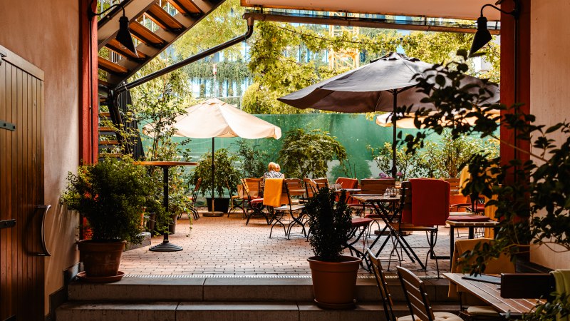 Ein gemütlicher Innenhof eines Cafés mit Tischen, Stühlen und Sonnenschirmen, umgeben von grünen Pflanzen und Bäumen., © SMG_Sarah Schmid