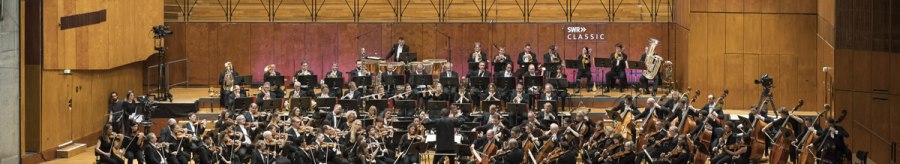 Das SWR Symphonieorchester spielt auf einer gro&szlig;en B&uuml;hne vor Publikum. Die Musiker tragen formelle Kleidung, und der Dirigent steht in der Mitte., &copy; SWR Alexander Kluge
