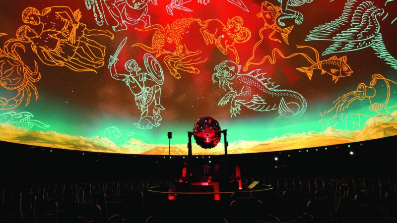 Ein Planetarium zeigt leuchtende Sternbilder an der Kuppel. Ein zentraler Sternenprojektor beleuchtet die Szenerie, während die Sitzreihen leer sind., © Planetarium Stuttgart