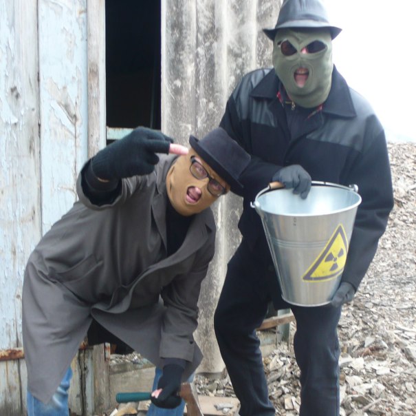 Zwei maskierte Personen in M&auml;nteln und H&uuml;ten posieren vor einem alten Geb&auml;ude. Eine Person h&auml;lt einen Eimer mit einem Warnsymbol., &copy; Die Alten