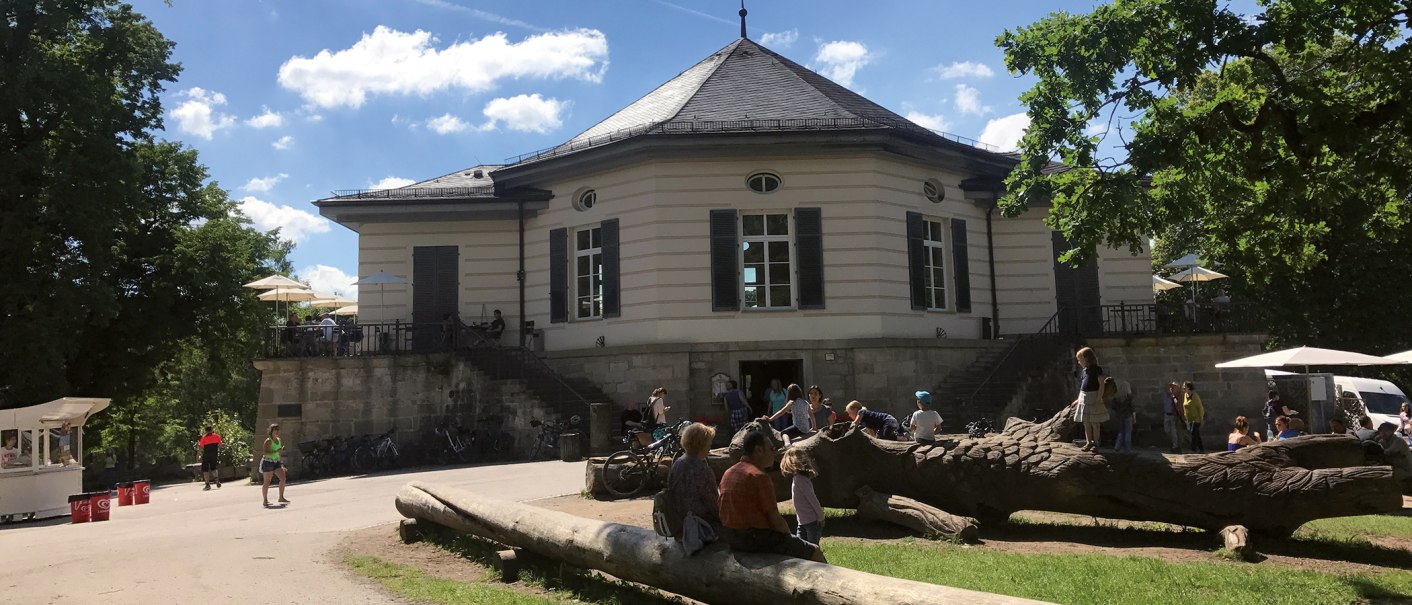 Das Bärenschlössle in Stuttgart, umgeben von Bäumen, mit Menschen, die draußen bei sonnigem Wetter entspannen und spielen., © SMG