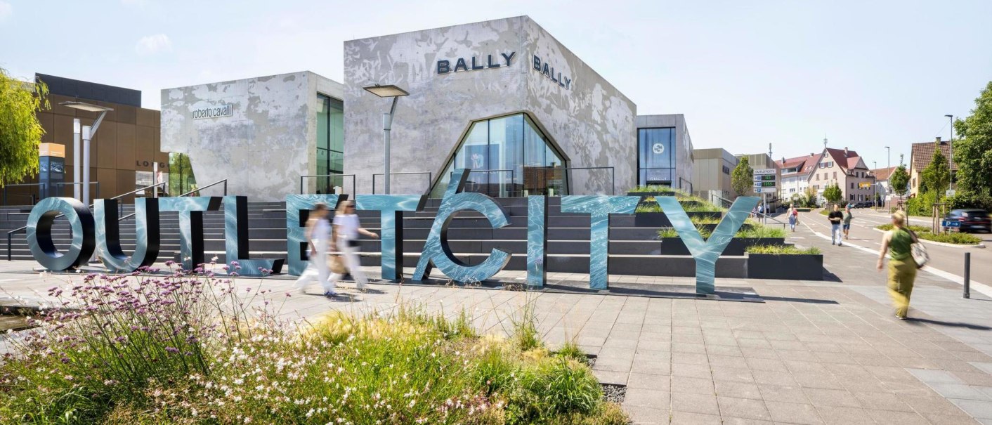Large letters spell out the word 'OUTLETCITY' in front of modern buildings. People walk by while flowers bloom in the foreground., &copy; OUTLETCITY AG