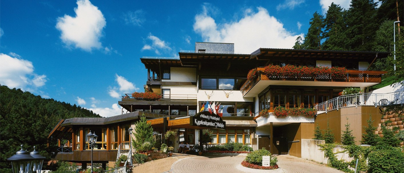 Hotel building with flower balconies, surrounded by forest and under a blue sky. A paved path leads to the entrance., © TOMAS