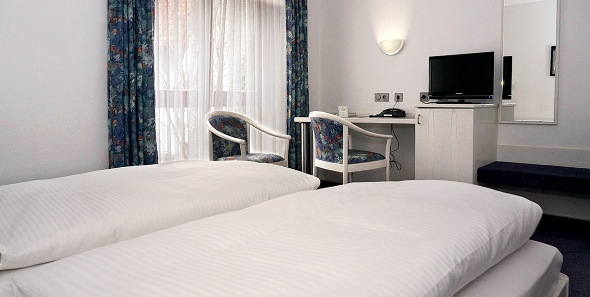 Ein schlichtes Hotelzimmer mit zwei Einzelbetten, einem Schreibtisch mit Stühlen, einem Fernseher und einem Fenster mit Vorhängen., © TOMAS Ein schlichtes Hotelzimmer mit zwei Einzelbetten, einem Schreibtisch mit Stühlen, einem Fernseher und einem Fenster mit Vorhängen., © TOMAS