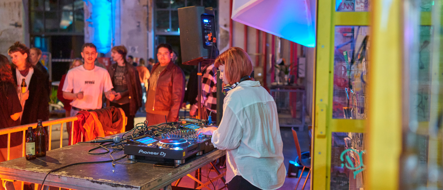 A DJ plays at a party, surrounded by dancing guests. The scene is colorfully lit and there is a lively atmosphere., &copy; PV Projekt Verlag GmbH