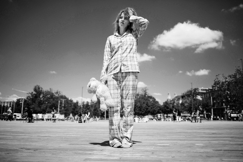 Eine Frau in kariertem Pyjama steht auf einem gro&szlig;en Platz und h&auml;lt ein Pl&uuml;schtier. Der Himmel ist klar mit einigen Wolken, und im Hintergrund sind B&auml;ume und Geb&auml;ude zu sehen., &copy; Rosenau Kultur e.V.