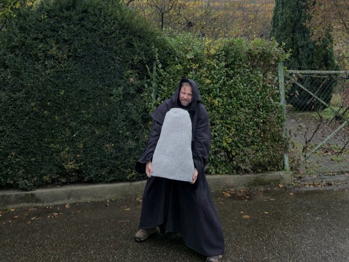 Ein Mann in einem dunklen Kostüm hält einen großen, grauen Stein. Er steht auf einer nassen Straße vor einer Hecke., © Cool-Tours StattReisen Ein Mann in einem dunklen Kostüm hält einen großen, grauen Stein. Er steht auf einer nassen Straße vor einer Hecke., © Cool-Tours StattReisen