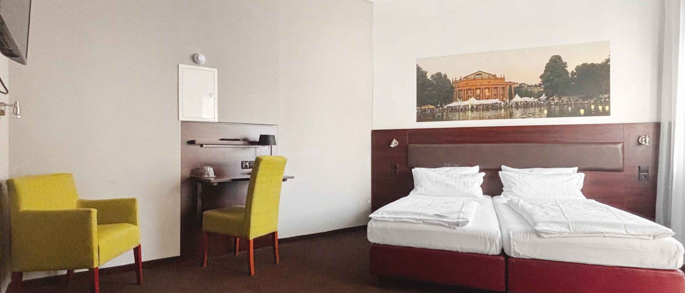 Ein Hotelzimmer mit zwei Einzelbetten, einem gelben Sessel und einem Schreibtisch. An der Wand h&auml;ngt ein Bild eines Geb&auml;udes., &copy; Novum Hospitality