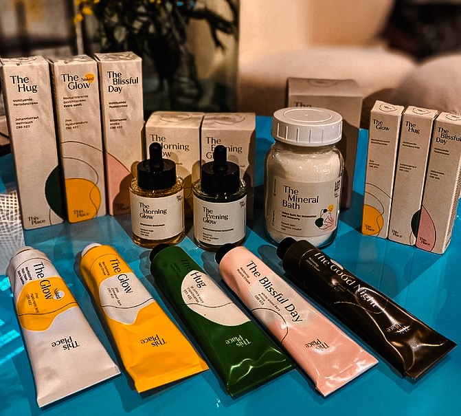 Kosmetikprodukte auf einem blauen Tisch, darunter Tuben und Flaschen mit Aufschriften wie 'The Glow' und 'The Mineral Bath', umgeben von gelben Blumen., © Merz & Benzing GmbH