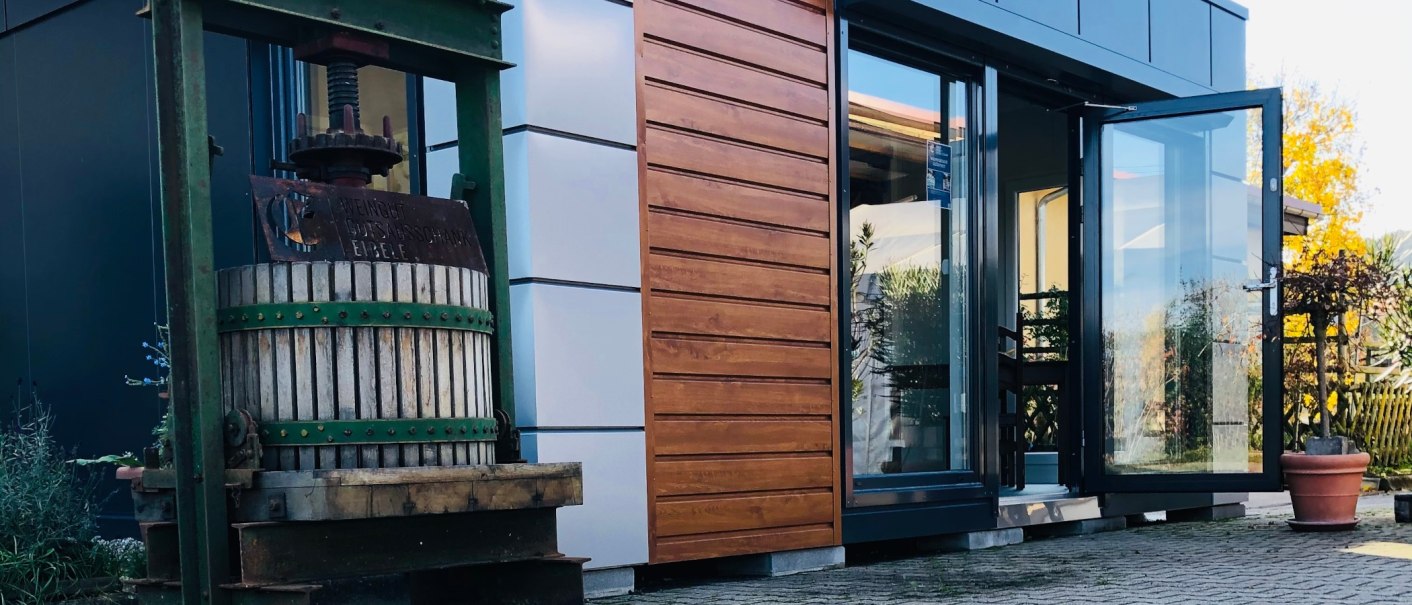 An old wine press stands in front of a modern building with wooden cladding and a glass front. Plants and a sunny sky can be seen in the background., &copy; Weingut Ei&szlig;ele