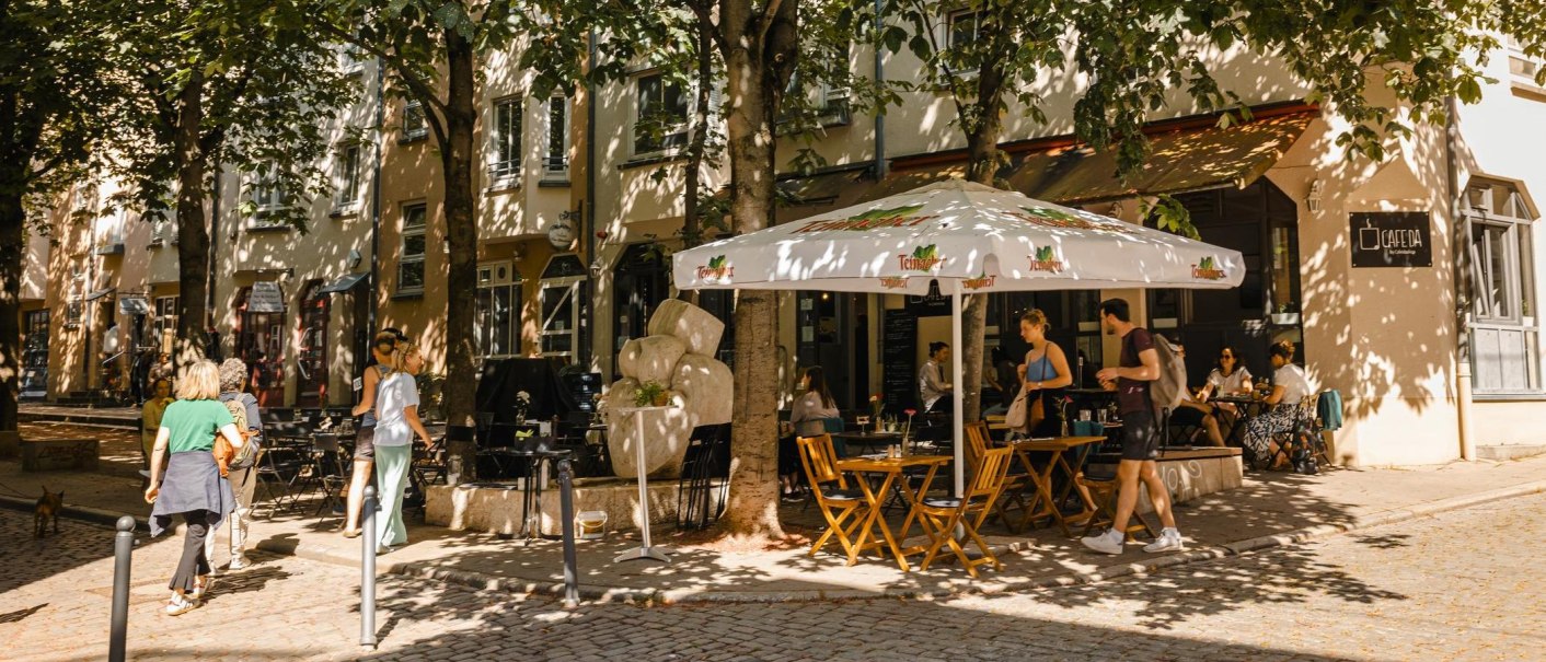 Ein gemütliches Straßencafé mit Sonnenschirm und Holztischen. Menschen sitzen und gehen vorbei, umgeben von Bäumen und Schatten., © Stuttgart Marketing GmbH - Sarah Schmid