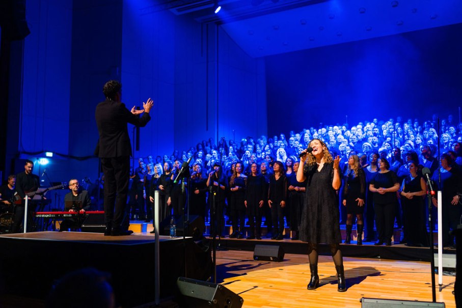 Ein großer Chor singt auf einer Bühne, beleuchtet von blauem Licht. Eine Solistin steht vorne, während ein Dirigent den Chor leitet., © Ludmilla Parsyak Ein großer Chor singt auf einer Bühne, beleuchtet von blauem Licht. Eine Solistin steht vorne, während ein Dirigent den Chor leitet., © Ludmilla Parsyak