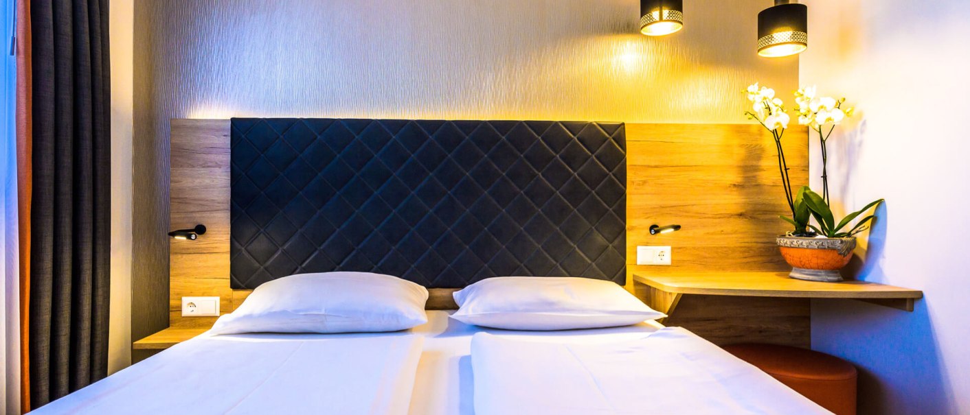 Modern double room with large bed, two pillows, bedside lamps and orchid on wooden table., © PLAZA INN Rieker Stuttgart Hauptbahnhof