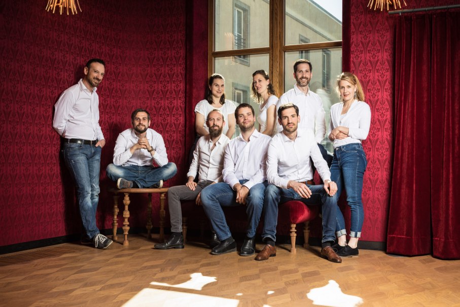 Neun Personen in weißer Kleidung posieren in einem stilvollen Raum mit roten Wänden und einem großen Fenster. Sie wirken entspannt und freundlich., © Voces Suaves Neun Personen in weißer Kleidung posieren in einem stilvollen Raum mit roten Wänden und einem großen Fenster. Sie wirken entspannt und freundlich., © Voces Suaves