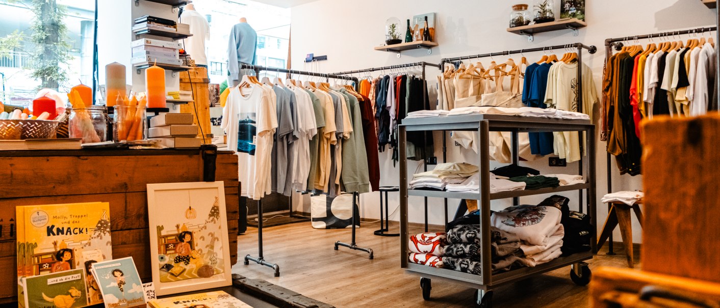 Ein Bekleidungsgeschäft mit T-Shirts auf Kleiderstangen, Regalen mit Kleidung und Dekorationen wie Kerzen und Büchern., © SMG_Sarah Schmid