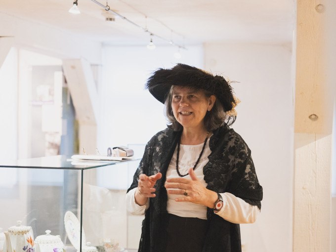 Eine Frau in historischer Kleidung steht in einem Museum neben einer Vitrine mit Porzellan. Sie trägt einen schwarzen Hut und eine Spitzenjacke., © Eigenbetrieb Tourismus und Citymanagement Schorndorf Eine Frau in historischer Kleidung steht in einem Museum neben einer Vitrine mit Porzellan. Sie trägt einen schwarzen Hut und eine Spitzenjacke., © Eigenbetrieb Tourismus und Citymanagement Schorndorf