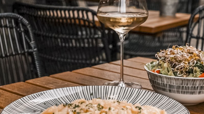 A glass of white wine, a plate of pasta and a salad on a wooden table in a restaurant with woven chairs., © SMG, itchyfeet-travel.de A glass of white wine, a plate of pasta and a salad on a wooden table in a restaurant with woven chairs., © SMG, itchyfeet-travel.de