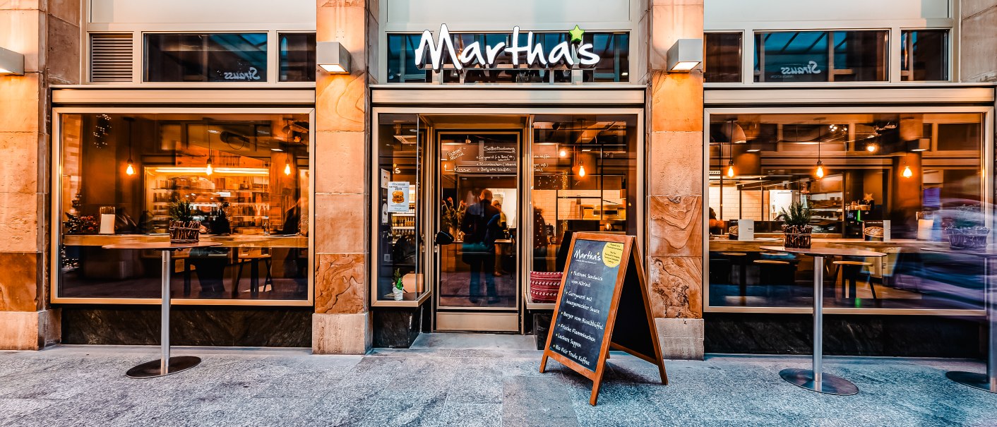 Eingang von Martha's im Königsbau mit großen Fenstern, Tischen und einer Tafel im Freien. Innenbeleuchtung und Dekor sind sichtbar., © Andreas Dalferth Eingang von Martha's im Königsbau mit großen Fenstern, Tischen und einer Tafel im Freien. Innenbeleuchtung und Dekor sind sichtbar., © Andreas Dalferth