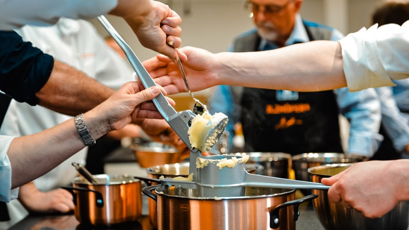 Mehrere Personen drücken Kartoffeln durch eine Presse in einen Topf. Im Hintergrund sind weitere Töpfe und eine Person mit Schürze zu sehen., © ILZHÖFER´s Event-Kochschule Mehrere Personen drücken Kartoffeln durch eine Presse in einen Topf. Im Hintergrund sind weitere Töpfe und eine Person mit Schürze zu sehen., © ILZHÖFER´s Event-Kochschule