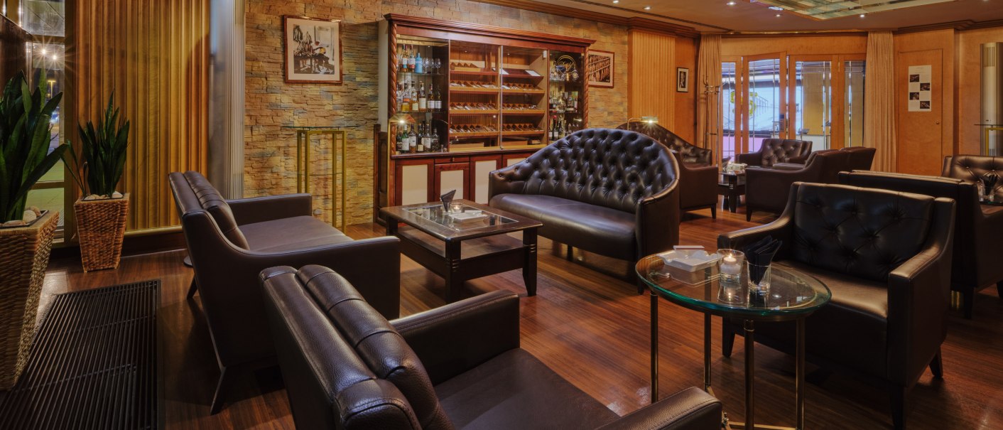 Elegant lounge with leather armchairs, wooden floor, plants and a humidor in the background. Subtle lighting and stylish furnishings., © Steigenberger Graf Zeppelin