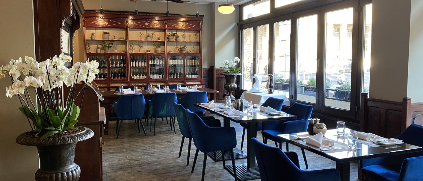 Elegantes Restaurant mit blauen Stühlen, Holzboden und großen Fenstern. Orchideen und eine Vitrine mit Flaschen schmücken den Raum., © Steigenberger Hotels GmbH