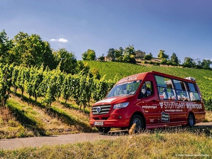 A red tour bus with the inscription 'Stuttgart Weintour' drives through green vineyards under a blue sky., &copy; Willms Touristik