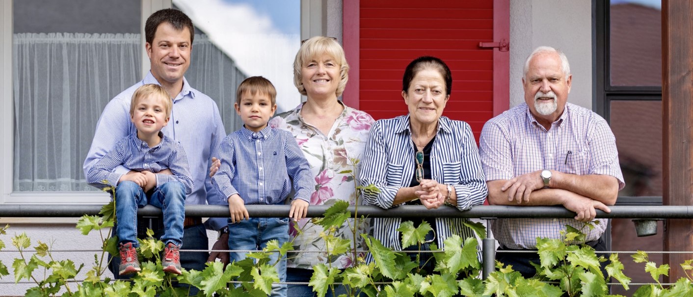 A smiling family stands on a balcony. Two children sit on the railing, surrounded by adults. Green plants can be seen in the foreground., © Weingut Zaiß