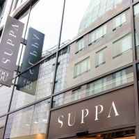 Glass fa&ccedil;ade of a modern building with several SUPPA signs. The reflection shows the surrounding architecture., &copy; Sarah Schmid