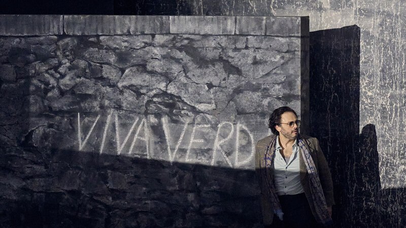 Ein Mann lehnt an einer grauen Steinwand mit der Aufschrift 'VIVA VERDI'. Die Szene wirkt dramatisch und düster., © Württembergische Staatstheater Stuttgart Ein Mann lehnt an einer grauen Steinwand mit der Aufschrift 'VIVA VERDI'. Die Szene wirkt dramatisch und düster., © Württembergische Staatstheater Stuttgart