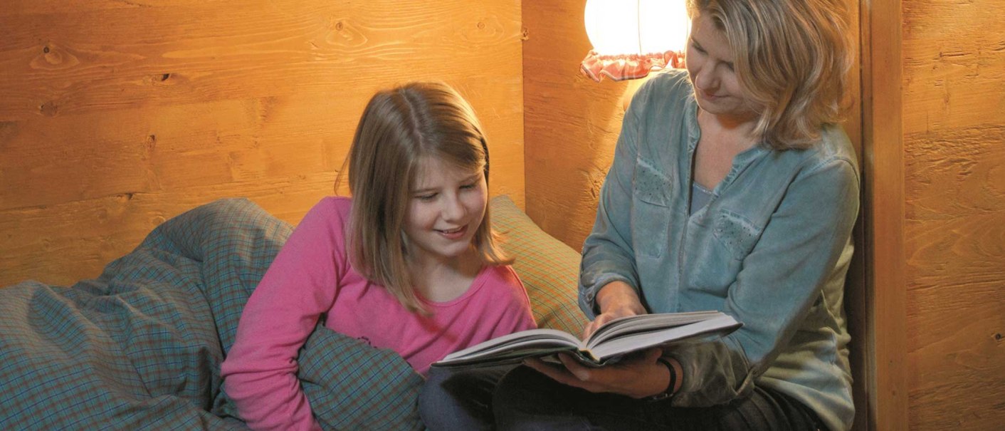 A woman is reading a book to a girl in a cozy wooden room. Both look relaxed and are smiling., &copy; Tripsdrill Natur-Resort GmbH