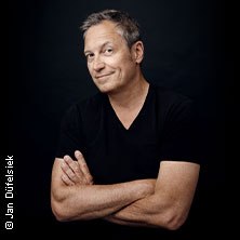 A man in a black T-shirt stands with his arms folded in front of a dark background and smiles slightly., &copy; see on the left