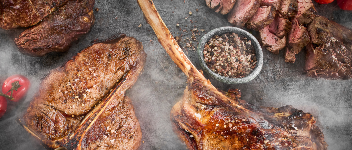 Grilled steaks on a table, surrounded by spices and tomatoes. The steaks are juicy and sprinkled with salt, next to a bowl of peppercorns., © MAREDO Grilled steaks on a table, surrounded by spices and tomatoes. The steaks are juicy and sprinkled with salt, next to a bowl of peppercorns., © MAREDO
