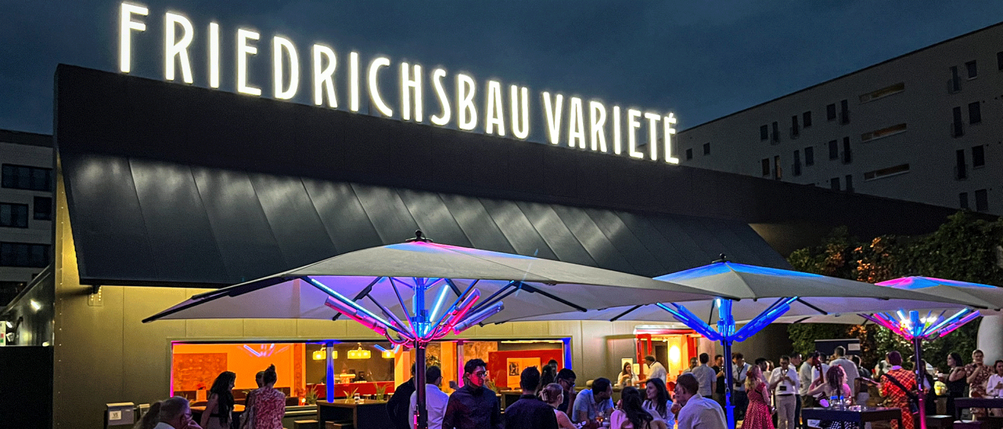 Abendliche Szene vor dem Friedrichsbau Varieté mit beleuchteten Sonnenschirmen und Menschen, die sich unterhalten und entspannen., © Friedrichsbau Varieté