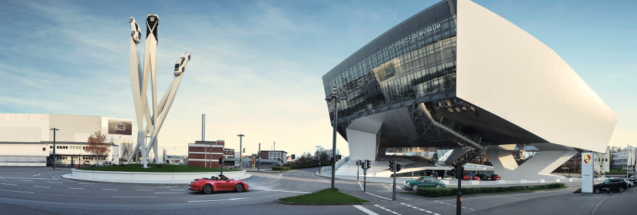 The Porsche Museum with its modern architecture and a sculpture of three Porsche cars on high pillars in daylight., © Porsche AG