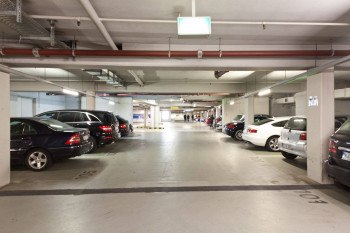 Underground parking garage with parked cars on both sides, bright lighting and visible pipes on the ceiling., © PLAZA INN Rieker Stuttgart Hauptbahnhof