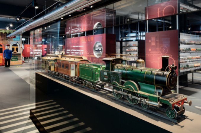 Historical model trains in a glass display case in the Märklineum. Further exhibits and information boards can be seen in the background., © Märklin Spielwaren Vertriebs-GmbH Historical model trains in a glass display case in the Märklineum. Further exhibits and information boards can be seen in the background., © Märklin Spielwaren Vertriebs-GmbH