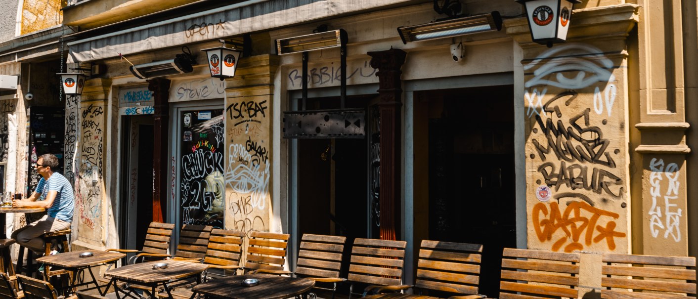 Bar La Concha mit Graffiti an der Fassade, leere Holztische davor, ein Mann sitzt draußen. Schilder von Erdinger Weißbräu und La Concha sichtbar., © Stuttgart-Marketing GmbH, Sarah Schmidt