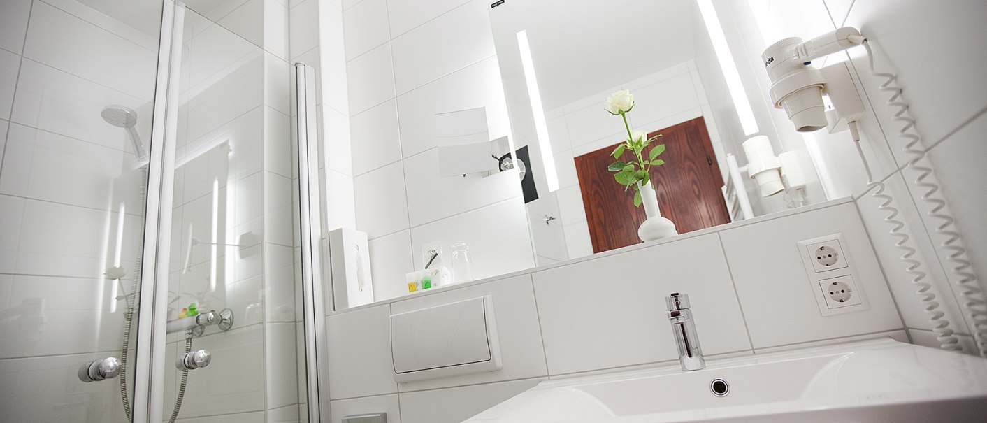 Modernes Badezimmer mit weißer Fliesenwand, Dusche, Waschbecken, Spiegel und einer weißen Blume in einer Vase., © Hotel Krone