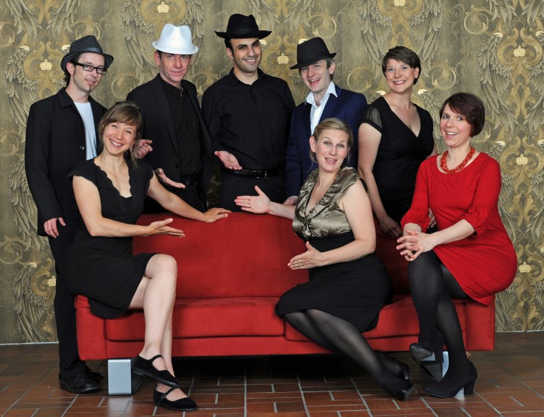 Acht Personen in eleganter Kleidung posieren um ein rotes Sofa vor einem kunstvollen Hintergrund. Sie l&auml;cheln und zeigen auf das Sofa., &copy; Yakup Zeyrek