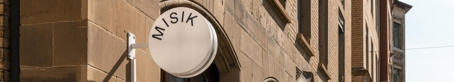Geb&auml;ude mit Sandsteinfassade, rundes Schild mit 'MISIK', Fenster mit Pflanzen, geschlossene Sonnenschirme auf der Stra&szlig;e., &copy; Stuttgart-Marketing GmbH, Sarah Schmid