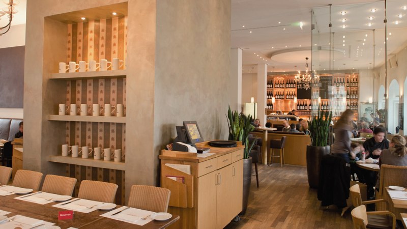 Stylish restaurant with wooden furniture, shelves full of cups and a cozy atmosphere. Guests sit at tables, with a bar in the background., © Aspectus Photographie Stuttgart