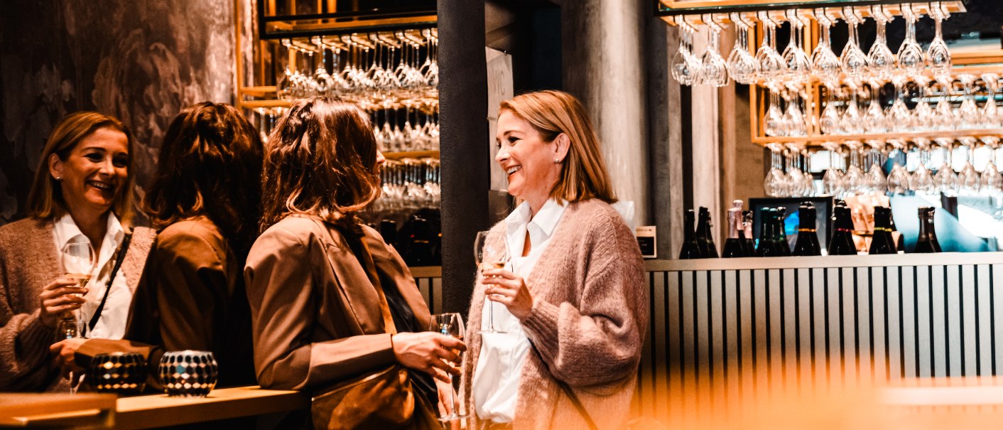 Drei Frauen unterhalten sich in einer stilvollen Bar. Sie halten Weingläser und lachen. Im Hintergrund sind viele hängende Weingläser zu sehen., © © Stuttgart-Marketing GmbH, wpsteinheisser