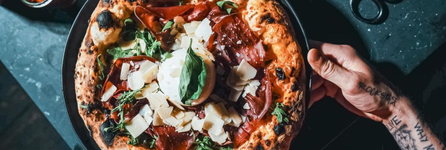 From the top: A pizza with a light, airy crust, ham, arugula, cheese, and basil on a black plate, held by a tattooed hand., &copy; L'Artista, Stuttgart
