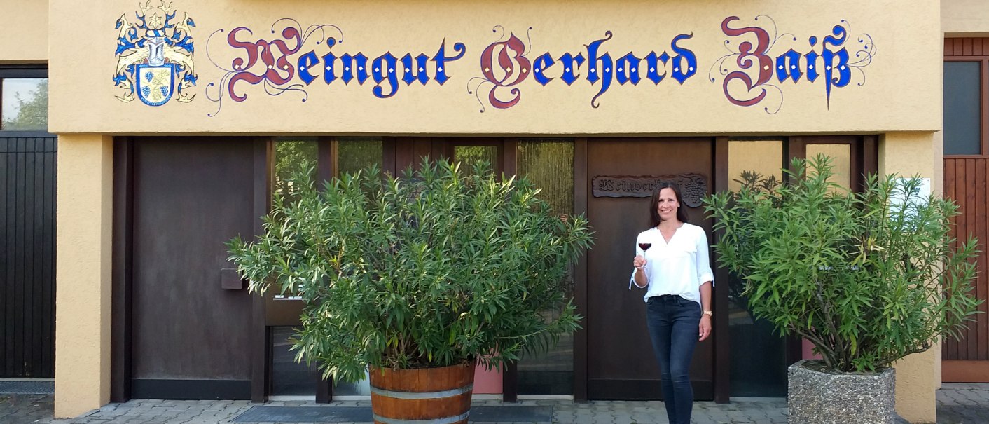Eine Frau steht vor dem Weingut Gerhard Zaiß, hält ein Glas Rotwein und lächelt. Das Gebäude hat eine dekorative Beschriftung und Pflanzen im Vordergrund., © Simone Mack