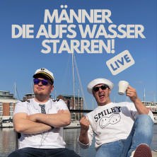 Zwei Männer sitzen entspannt vor einem Hafen. Einer trägt eine Kapitänsmütze, der andere hält eine Tasse. Im Hintergrund sind Boote und blauer Himmel zu sehen., © links im Bild Zwei Männer sitzen entspannt vor einem Hafen. Einer trägt eine Kapitänsmütze, der andere hält eine Tasse. Im Hintergrund sind Boote und blauer Himmel zu sehen., © links im Bild