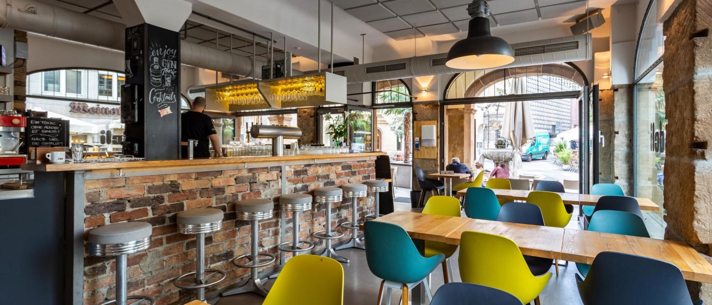 Modern café with a brick bar, colorful chairs and large windows. A man is standing behind the bar, passers-by can be seen outside., © deli, Stuttgart